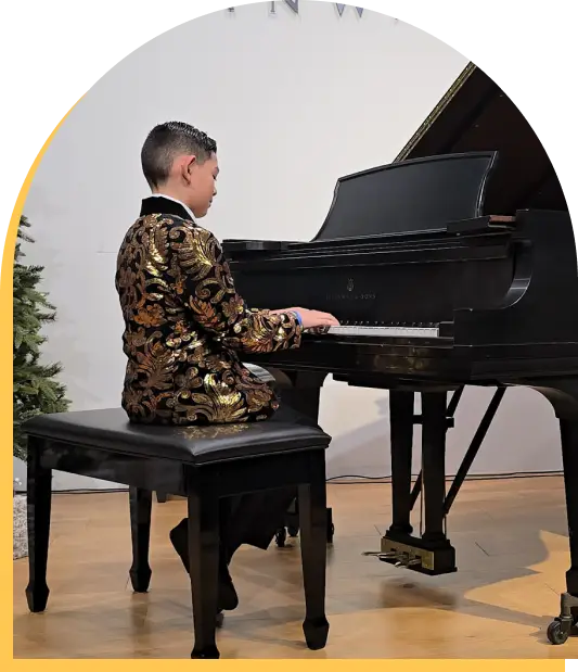 A young boy wearing a gold and black patterned jacket plays a grand piano on stage, seated on a black piano bench. A small tree is visible in the background—a testament to the talent fostered by music lessons Westchase has to offer.