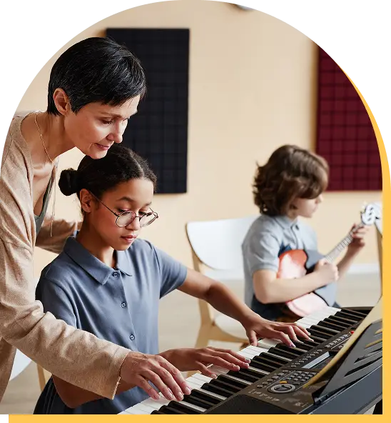 A woman guides a girl playing a keyboard, while a boy in the background plays an acoustic guitar. They are in a music classroom with soundproof panels on the walls, enjoying music lessons Westchase families love.
