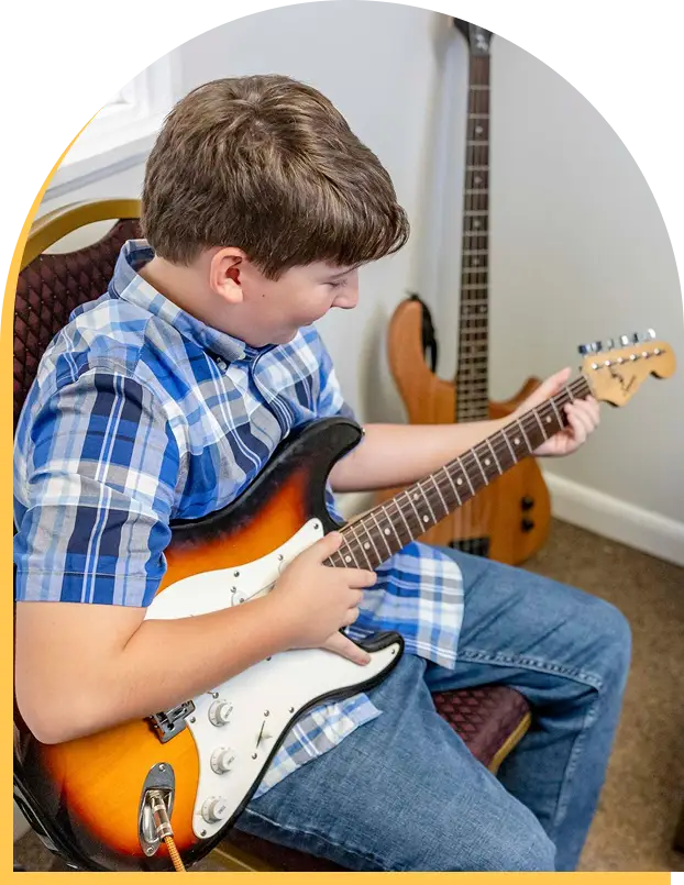 A boy in a blue plaid shirt and jeans sits on a chair playing a sunburst electric guitar, perhaps inspired by music lessons Westchase offers. A wooden bass guitar rests upright against the wall in the background.