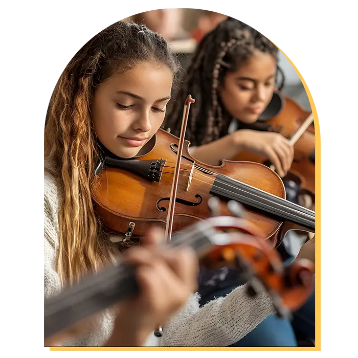 Two girls with long hair play violins in a classroom setting during music lessons Westchase, focused on their instruments. The foreground highlights one girl closely, while another plays behind her. Warm lighting creates a calm, studious atmosphere.