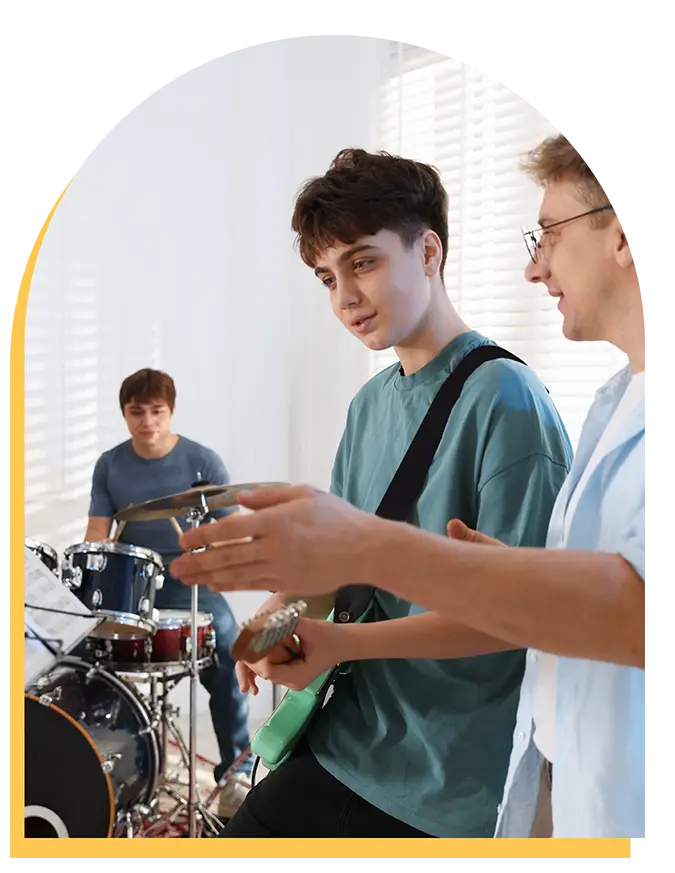 Three young people playing music together indoors; one plays drums in the background, while two stand in front, one holding a green electric guitar and the other gesturing as if explaining or discussing something.